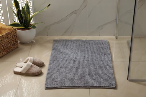 Szary dywanik łazienkowy, obok kapcie i prysznic